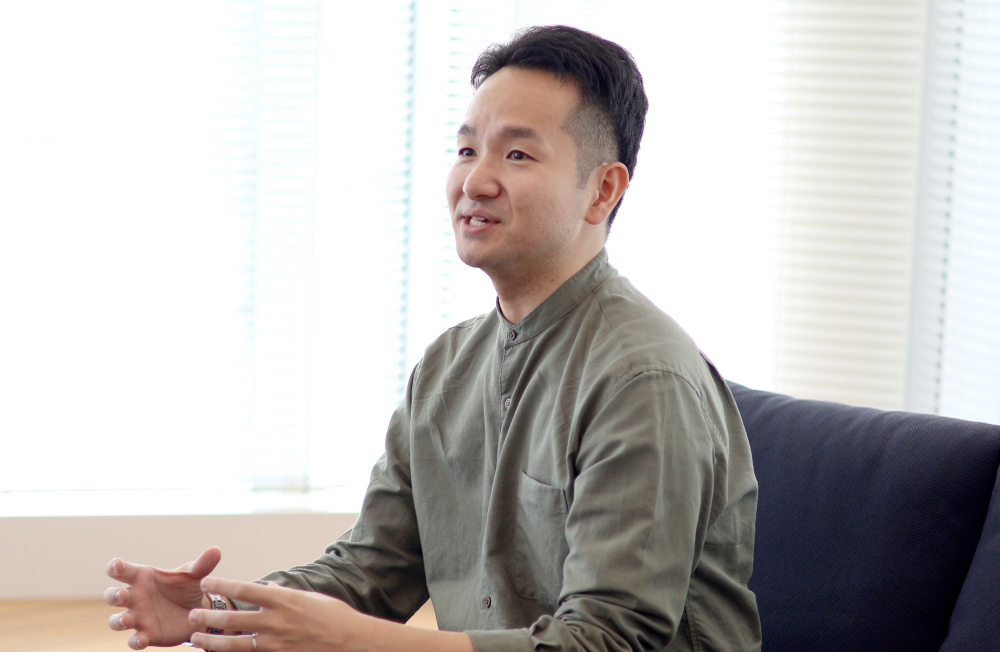 Ina speaking during an interview in a bright office. Sitting on a sofa, he gestures gently while talking with a calm smile. Natural light from the window creates an open and relaxed atmosphere.