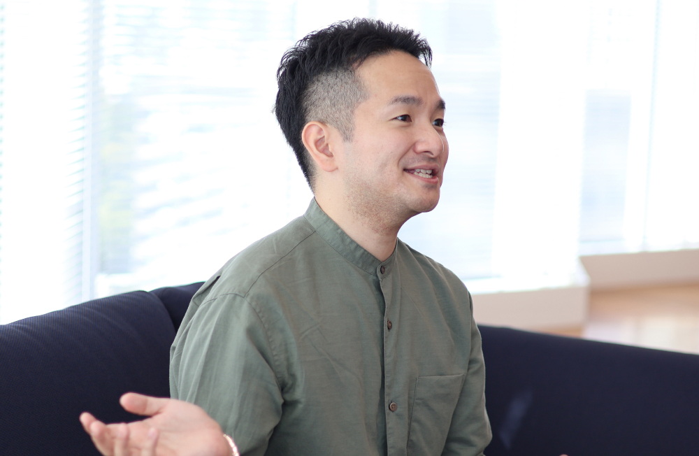Ina smiling during an interview. Wearing a green shirt, he sits on a sofa and gestures expressively while explaining his thoughts. His relaxed posture and cheerful expression convey a positive attitude.