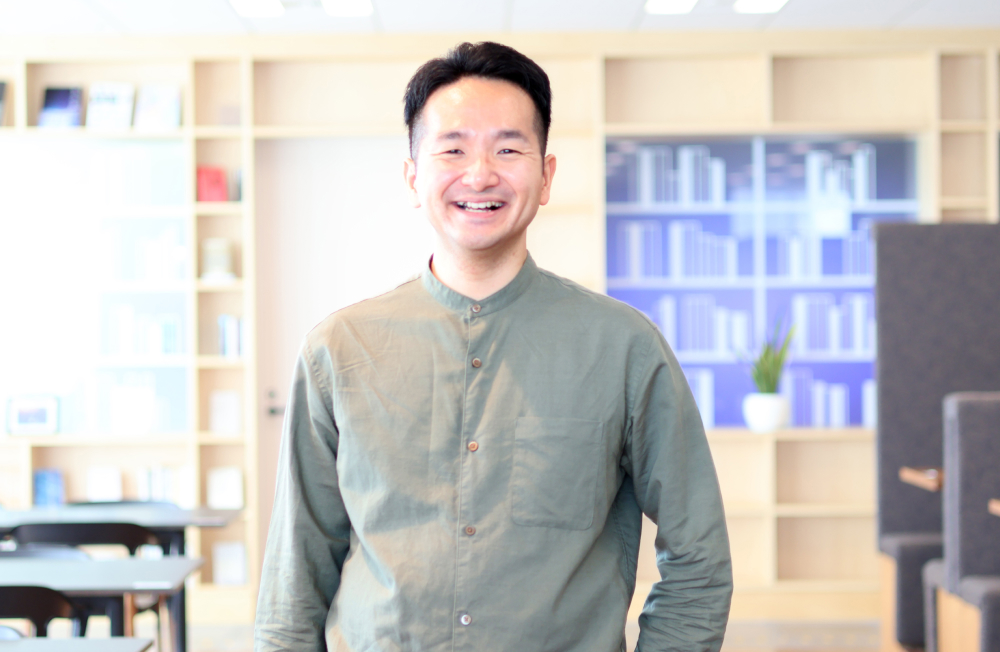 Ina smiling in the office. Bookshelves and plants in the background create a cozy, welcoming work environment. A natural moment captured after the interview.