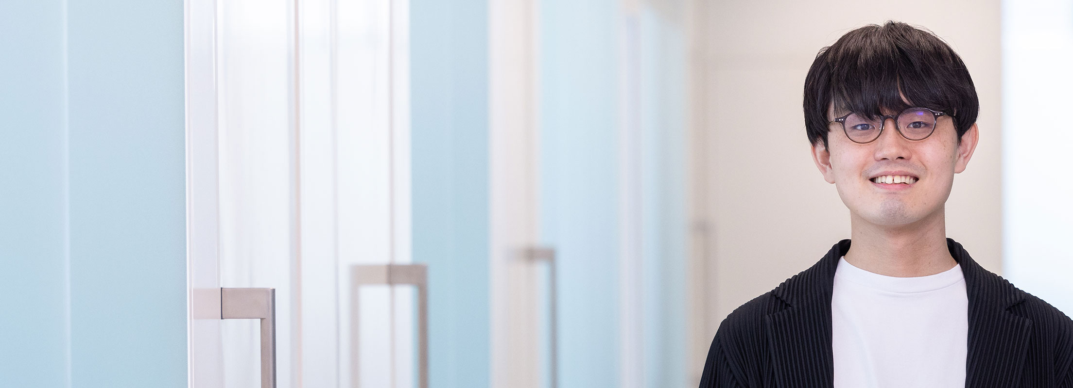 A man wearing glasses is smiling at the camera in a bright hallway.