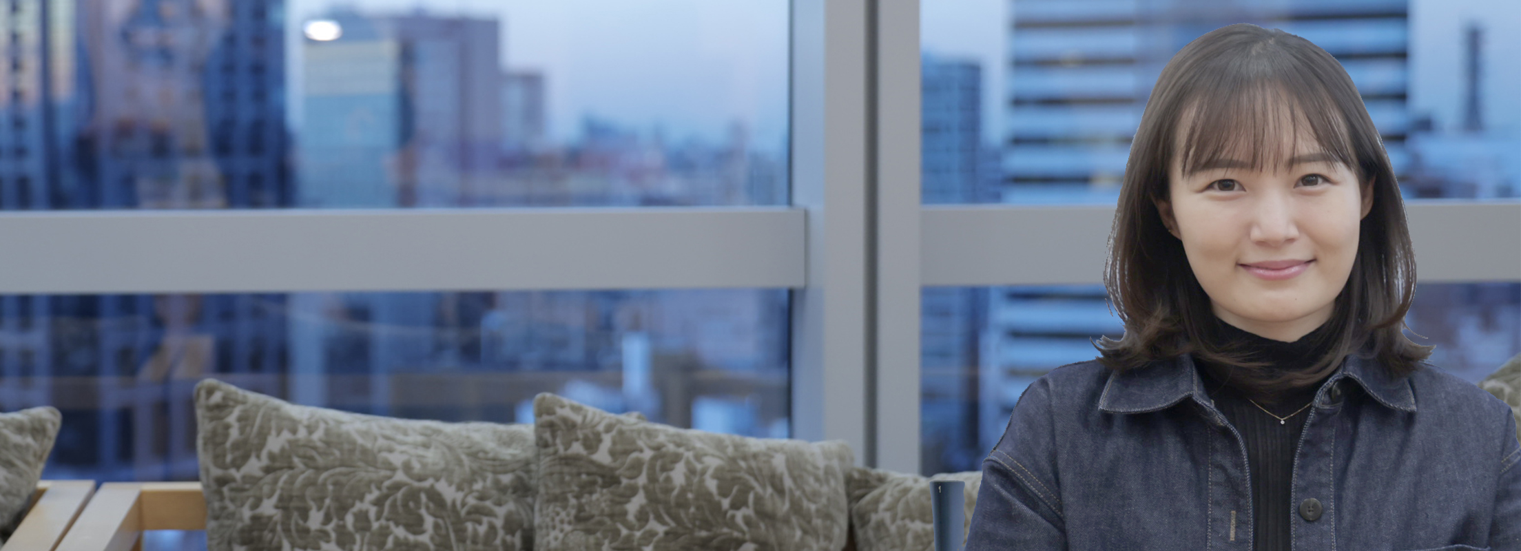 A woman is smiling at the camera indoors. In the background, there is a large window with a view of the cityscape.
