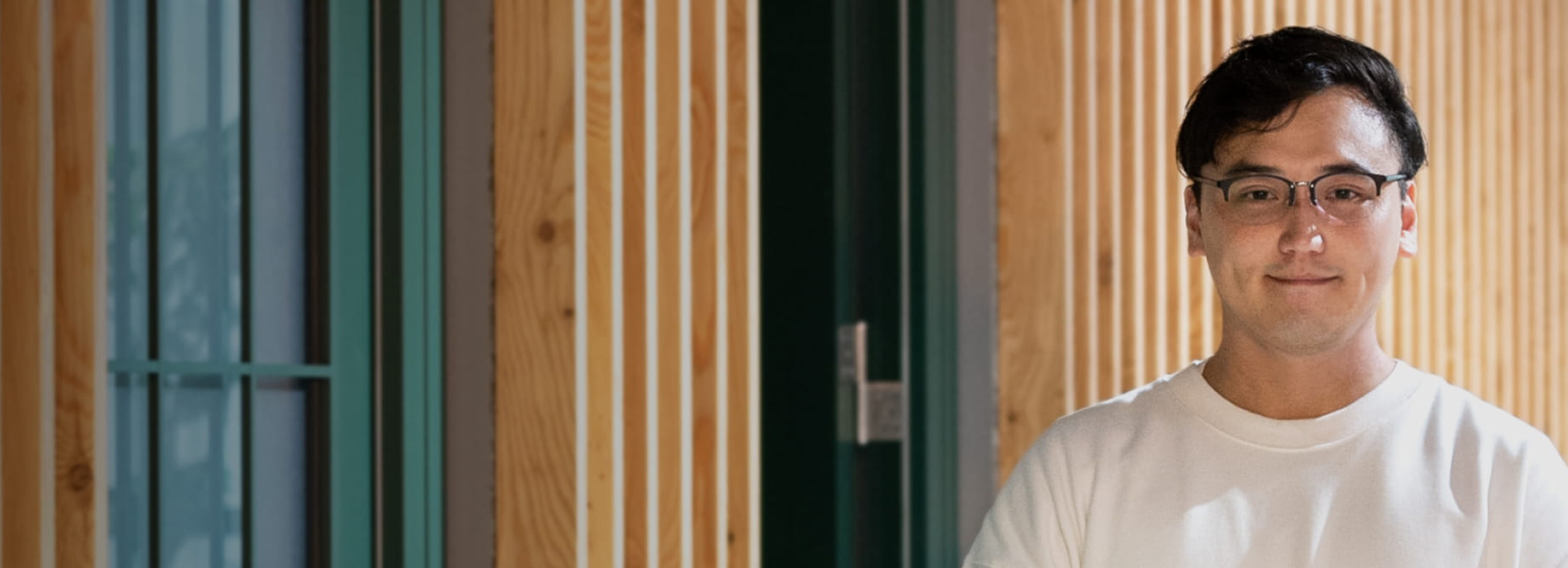 A man wearing glasses and a white shirt is smiling. In the background, there is a wooden wall and a window.