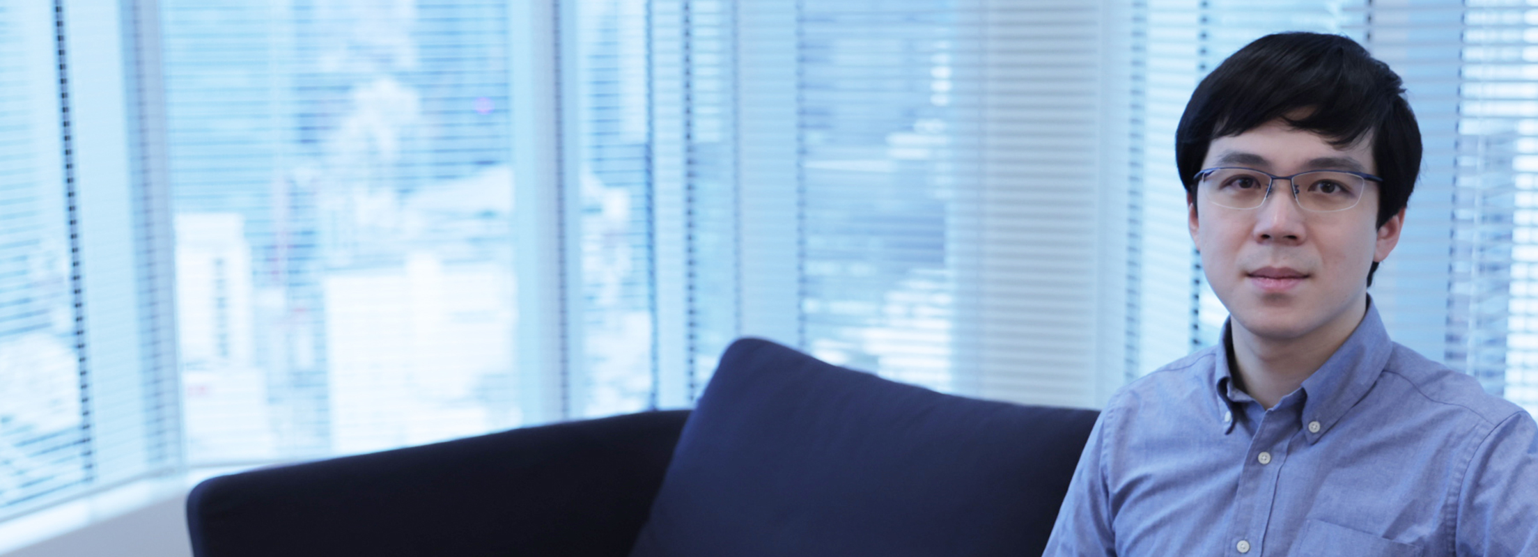 A man wearing glasses and a blue shirt is looking at the camera. In the background, there is a window and a sofa.