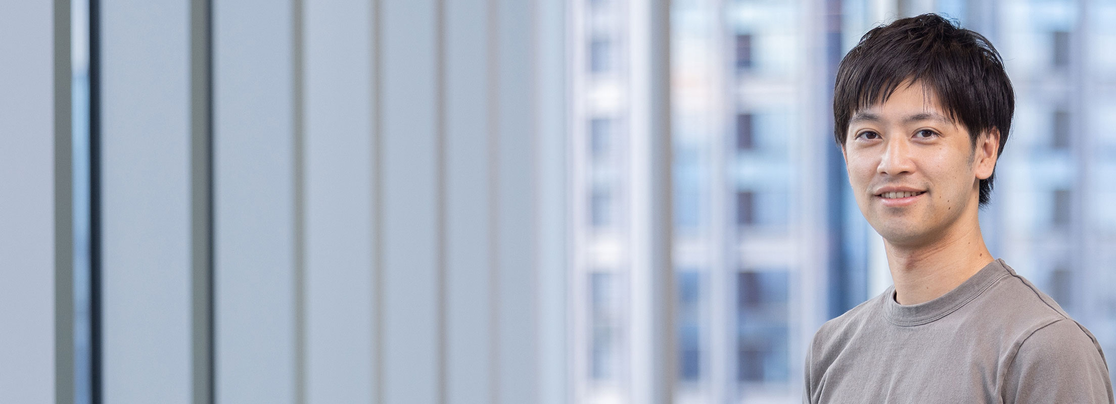 The photo shows a man in casual clothing smiling. In the background, there is a large window with a view of the cityscape.