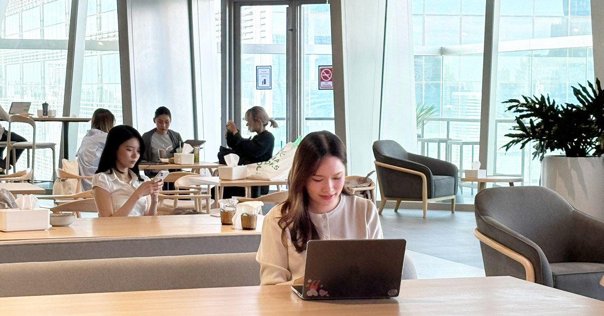 People are spending time in a café-style lounge. In the foreground, a person wearing a white long-sleeved top works on a laptop.