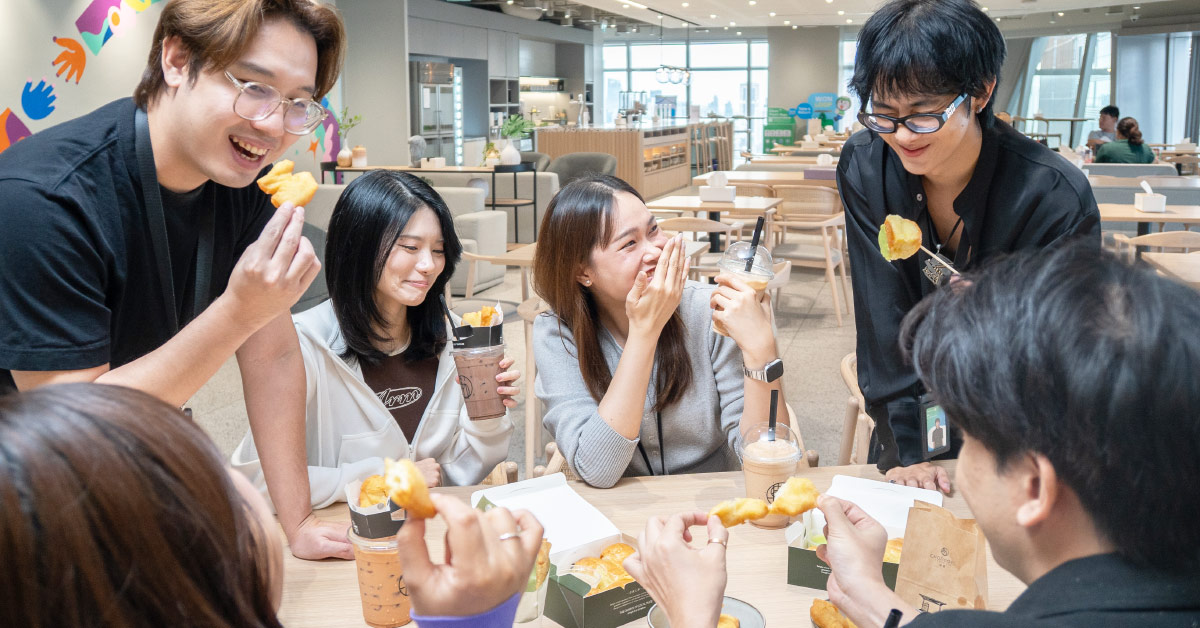 Six people gather around a table in a lounge, enjoying snacks and drinks while chatting.