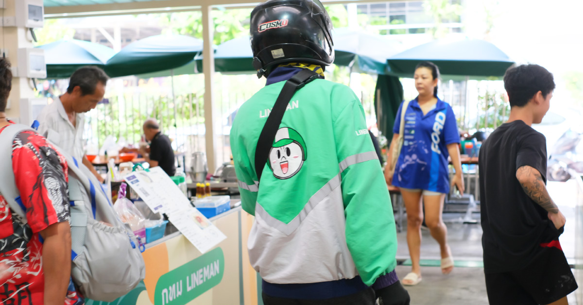 A LINE MAN delivery rider waits inside a restaurant to collect an order. Wearing a green-and-white delivery jacket and a helmet, the rider is seen from behind with the LINE MAN character printed on the back.