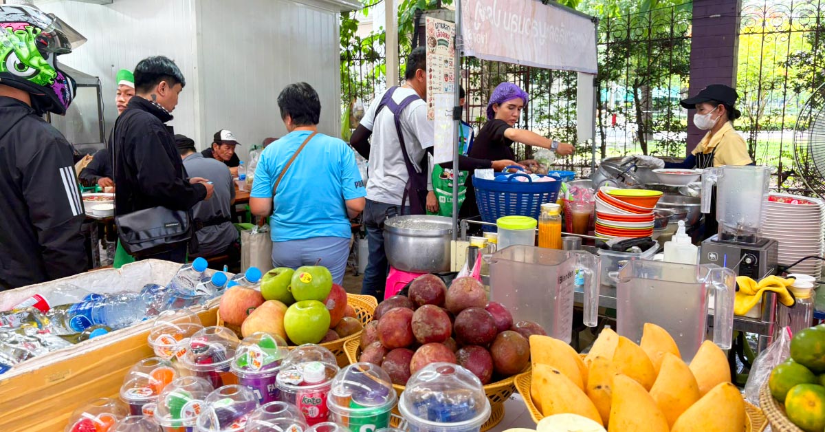 A busy outdoor food market filled with customers eating and shopping. Fresh fruit, bottled drinks, and cups of jelly desserts are displayed in the foreground, while vendors prepare food behind the counter.
