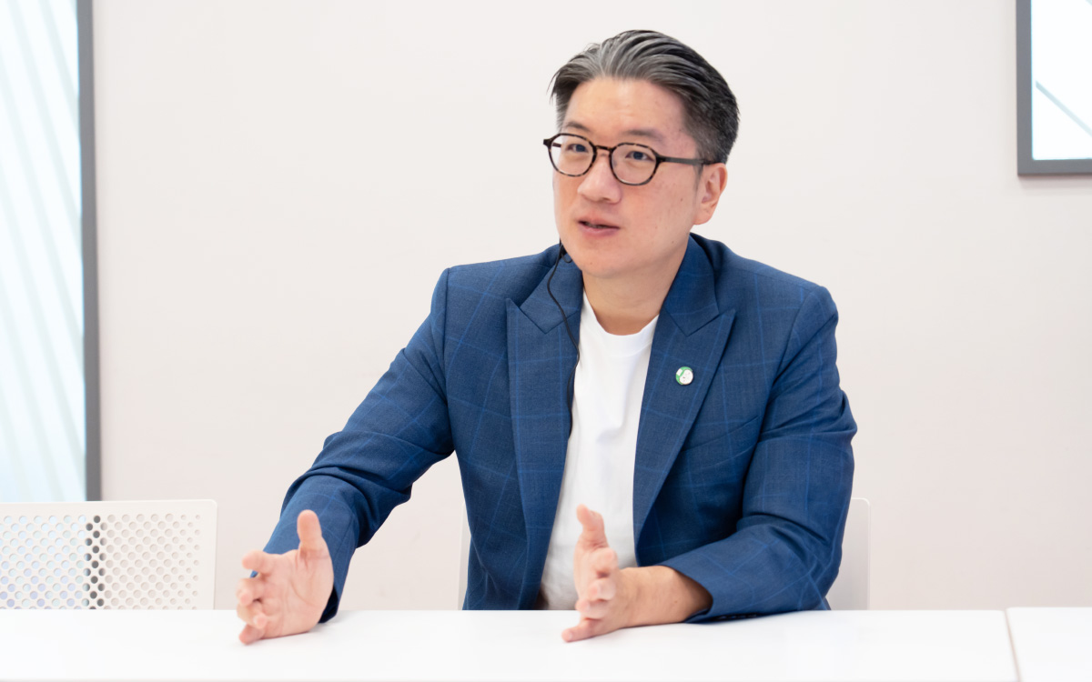 This is a bust shot of Yod Chinsupakul. His black hair, streaked with gray, is neatly combed back, and he is wearing round black-framed glasses. He is speaking with both hands extended slightly outward on the desk, and he is wearing an earpiece-style microphone. His expression is serious, and he appears to be explaining something while looking slightly to the right.