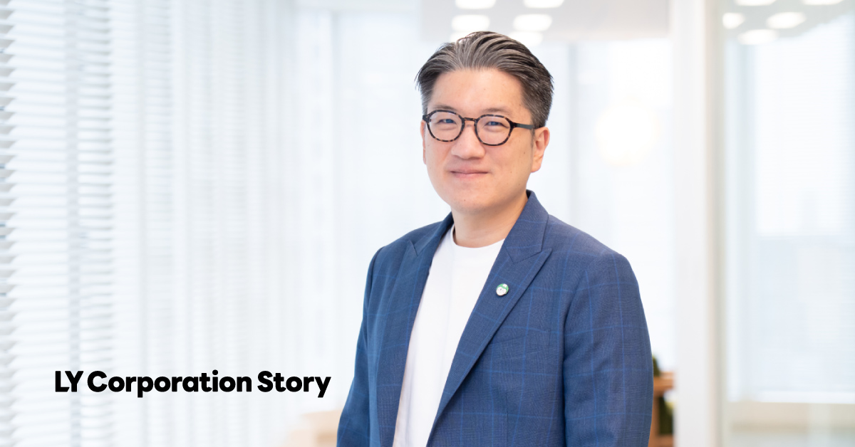 Yod Chinsupakul in a bust shot. His black hair, streaked with gray, is neatly combed back, and he is wearing round black-framed glasses. He has a gentle smile and is standing with his body slightly angled. The background shows a bright office space, and the text 