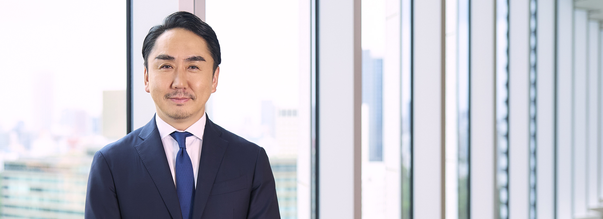 代表取締役社長 CEO（最高経営責任者）を務める出澤　剛の写真です。スーツを着た男性が、明るくモダンなオフィスで微笑んでいます。背景には大きな窓があります。