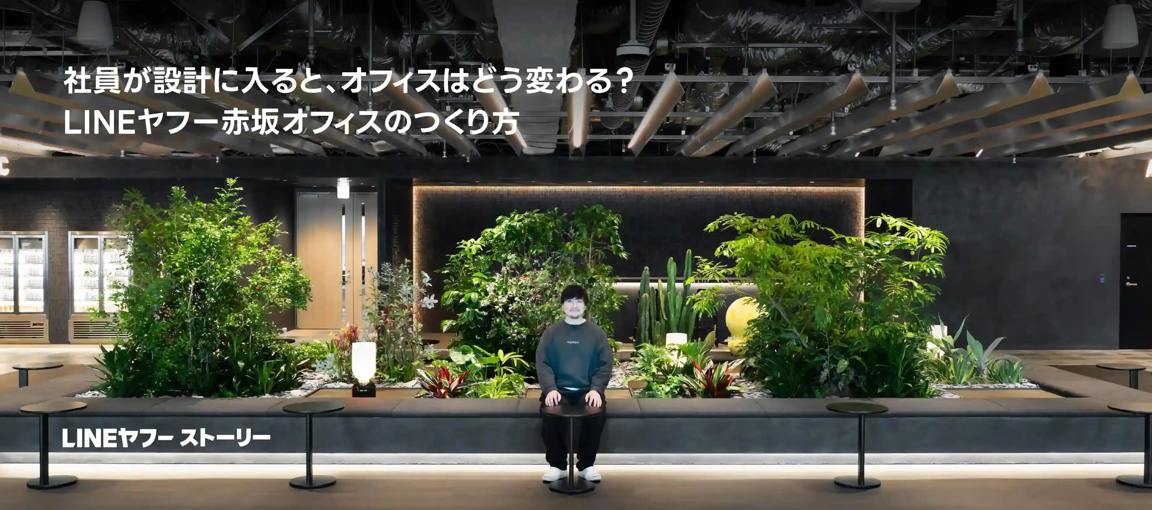 「社員が設計に入ると、オフィスはどう変わる？LINEヤフー赤坂オフィスのつくり方」という見出しのついた画像。薄暗くスタイリッシュな空間に、サボテンや観葉植物が豊富に配置された緑あふれるエリアがある。その手前の長い黒いベンチに、ダークグレーのトップスを着た男性が一人座っている。左下には「LINEヤフー ストーリー」の文字。