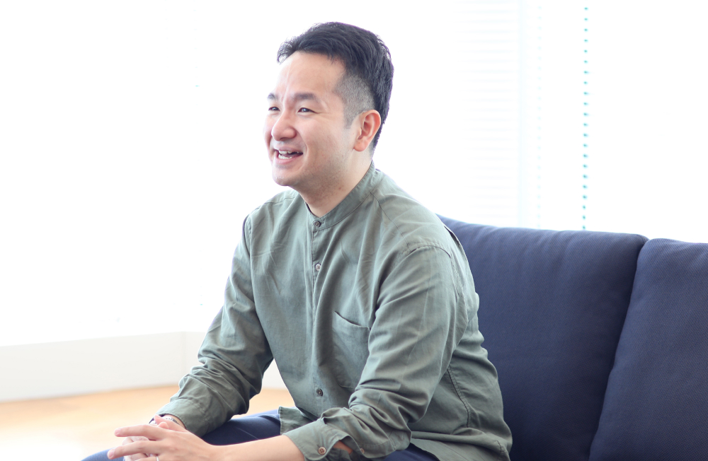 質問に答えながら笑顔を見せる伊奈さんのインタビュー風景。自然光が差し込む明るいオフィスで、真剣さと柔らかさが感じられる表情が印象的。