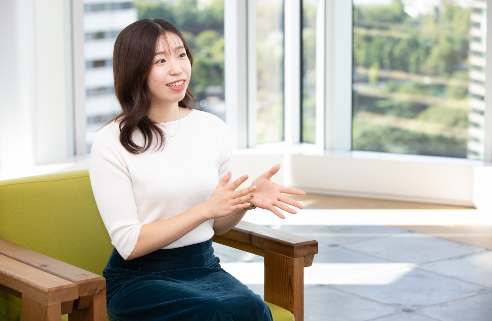 大きな窓の前でインタビューに答える出野さん。手振りを交えながら話している。自然光が差し込む明るいオフィスで、外には緑の多い風景が見える。