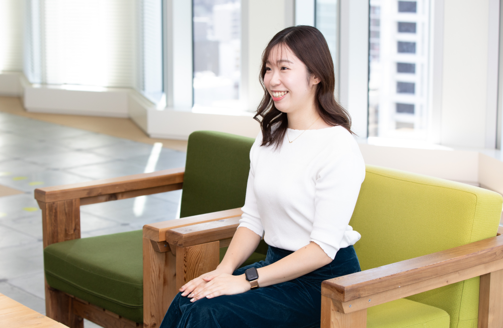 木製の肘掛けがついたソファに座り、穏やかに微笑む出野さん。白いトップスと濃紺のスカート姿で、左腕にスマートウォッチを着けている。背景には大きな窓が並び、都会的なビル群が見えるオフィス空間。