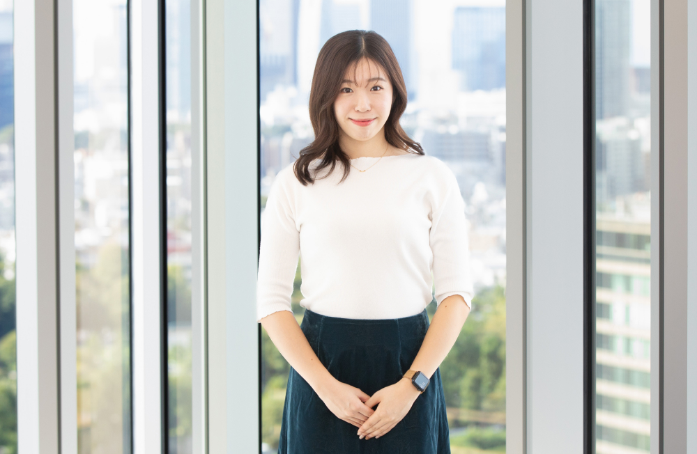 大きな窓の前で立ち、穏やかな笑顔でカメラを見つめる出野さん。白いニットとネイビーのスカートを着用し、両手を前で軽く組んでいる。背景には都会の景色と木々の緑が広がり、清潔感と落ち着いた印象を与える。