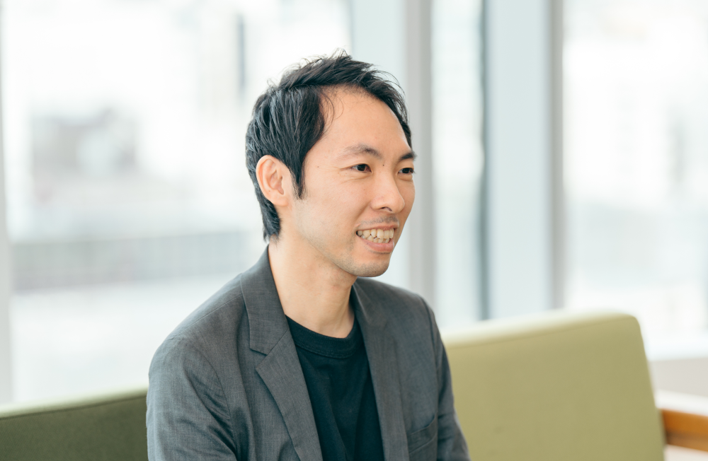 橋本 秀太郎さんのインタビュー記事の写真です。窓から自然光が差し込む中、ソファに座ったグレーのジャケット姿の橋本さんが、開放的な雰囲気の中で笑顔を見せながら会話している。