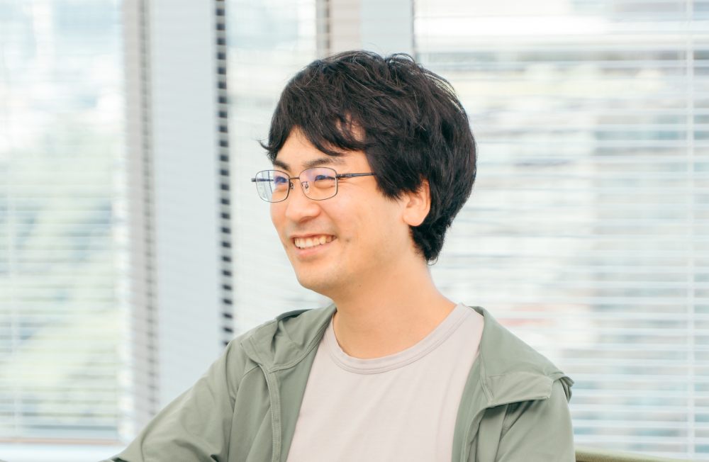 室内で椅子に座り、笑顔を見せている人物の上半身ショット。人物は黒髪で眼鏡をかけ、薄いグレーのTシャツに薄緑色のジップアップパーカーを羽織っている。背景にはブラインド越しに外の景色が見えている。