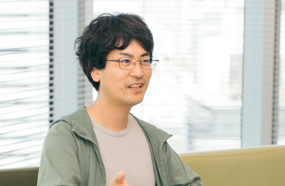 室内で椅子に座り、会話をしている様子の人物の上半身ショット。人物は黒髪で眼鏡をかけ、薄いグレーのTシャツの上に薄緑色のジップアップパーカーを羽織っている。口を少し開き、片手を前に出して話している仕草をしている。背景にはブラインドと外の景色が見えている。