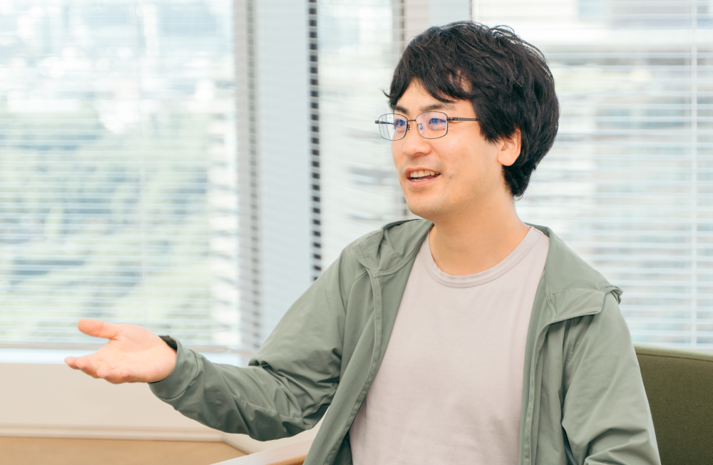室内で椅子に座り、会話の最中に片手を横に広げている人物の上半身ショット。人物は黒髪で眼鏡をかけ、薄いグレーのTシャツの上に薄緑色のジップアップパーカーを羽織っている。表情は明るく、話に熱を込めている様子が伝わる。背景にはブラインド越しに外の景色が見えている。