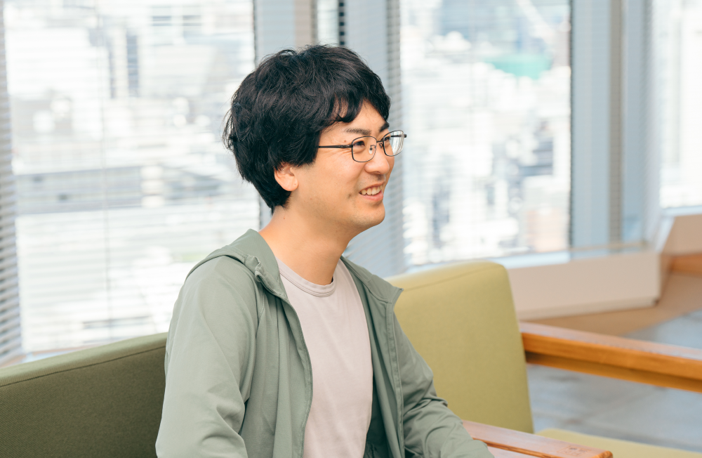 室内で椅子に座り、横を向いて笑顔を見せている人物の上半身ショット。人物は黒髪で眼鏡をかけ、薄いグレーのTシャツの上に薄緑色のジップアップパーカーを羽織っている。背景には窓から高層ビル群が見え、椅子の木製アームと緑色のクッションも写っている。