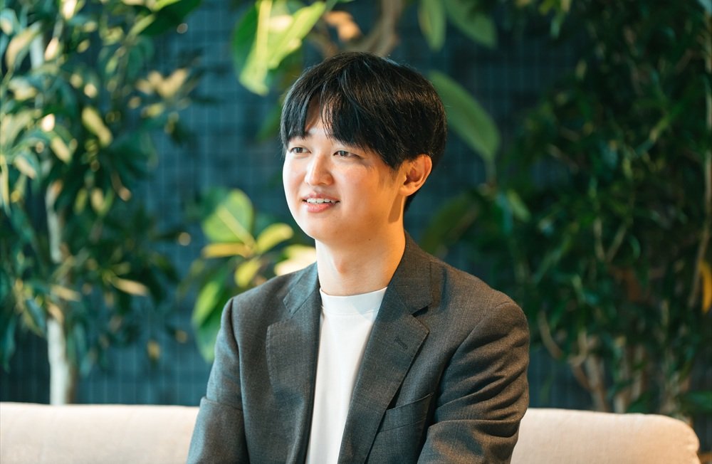 観葉植物を背景に、光安さんがソファに座って話している表情のカット。