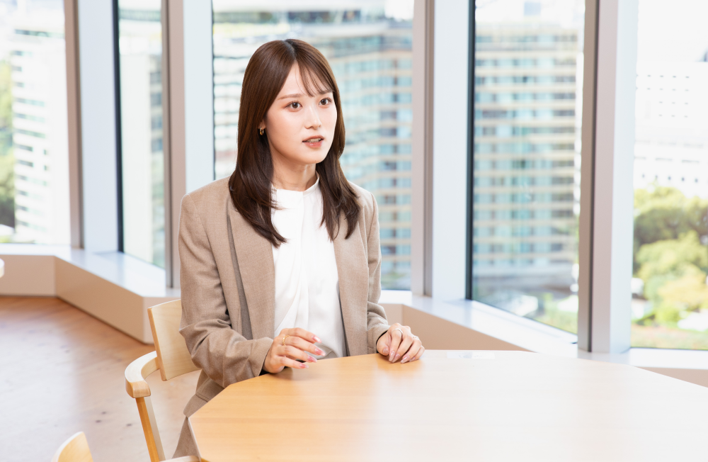 大きな窓のある明るいオフィスで、丸テーブルに座りインタビューに答える杉崎さん。白いブラウスにベージュのジャケットを着用し、両手をテーブルに添えて話をしている。背景には高層ビルや緑が見える落ち着いた空間。