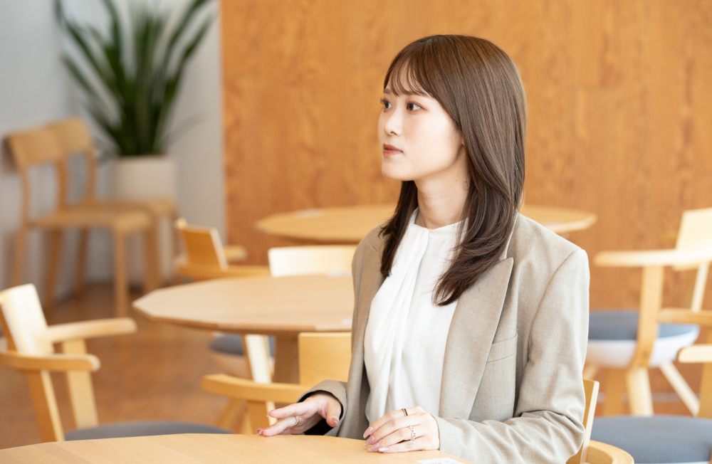 木目調の壁とナチュラルな木製家具に囲まれたカフェ風のスペースで、横を向いて話す杉崎さん。白いブラウスとベージュのスーツ姿で、真剣な表情をしている。奥には観葉植物と複数のテーブルと椅子が見える。