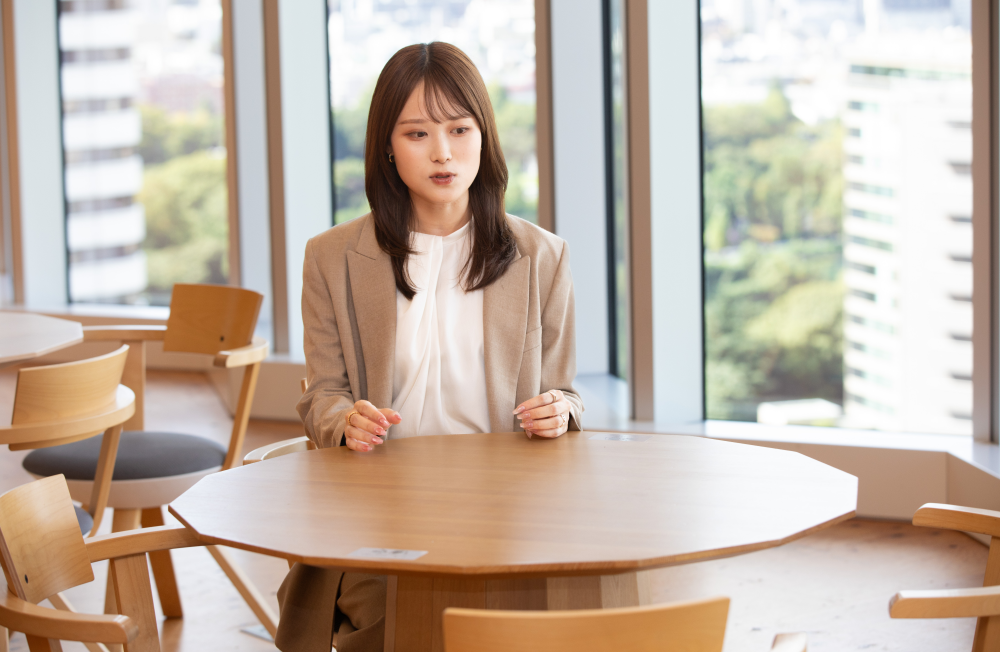 広い窓際のオフィスで、円形のテーブルに座り話している杉崎さん。白いブラウスにベージュのジャケットを着用し、両手を軽く握って前に出している。背景には木製の椅子が並び、窓の外には緑とビル群が広がる。