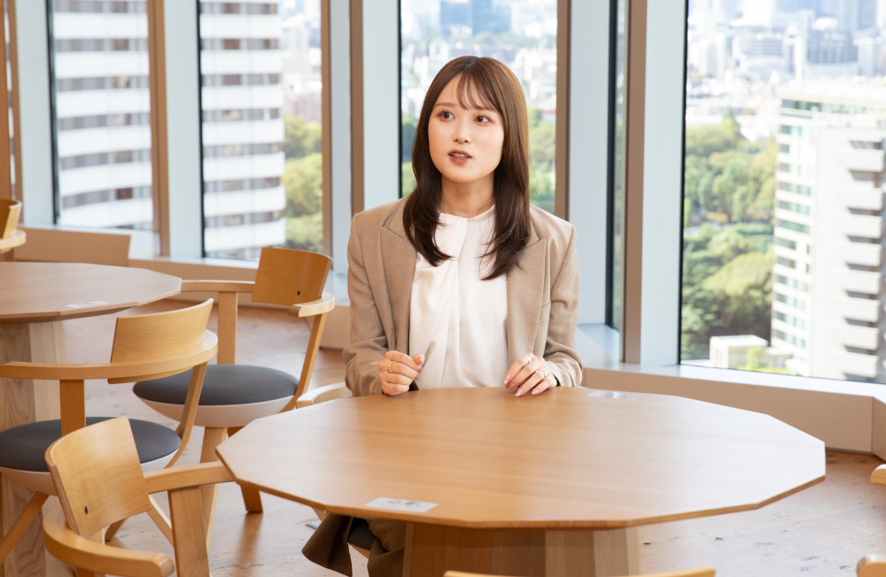 明るい自然光が差し込むオフィスで、テーブル越しにインタビューを受ける杉崎さん。白いインナーにベージュのジャケットを着用し、落ち着いた表情で話している。背景には大きな窓と都会の緑豊かな景観が見える。