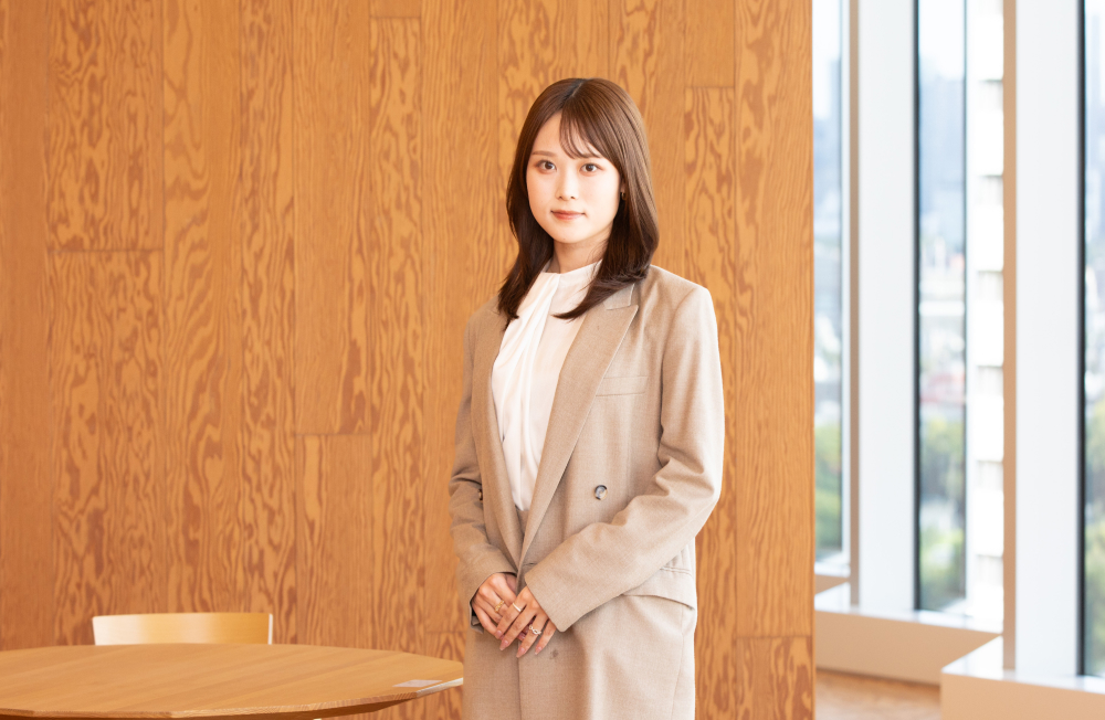 木目の壁の前に立ち、正面を向いて穏やかに微笑む杉崎さん。白いブラウスとベージュのスーツを着用し、両手を前で軽く組んでいる。背後には丸テーブルと窓が並び、明るく開放的なオフィス空間が広がる。
