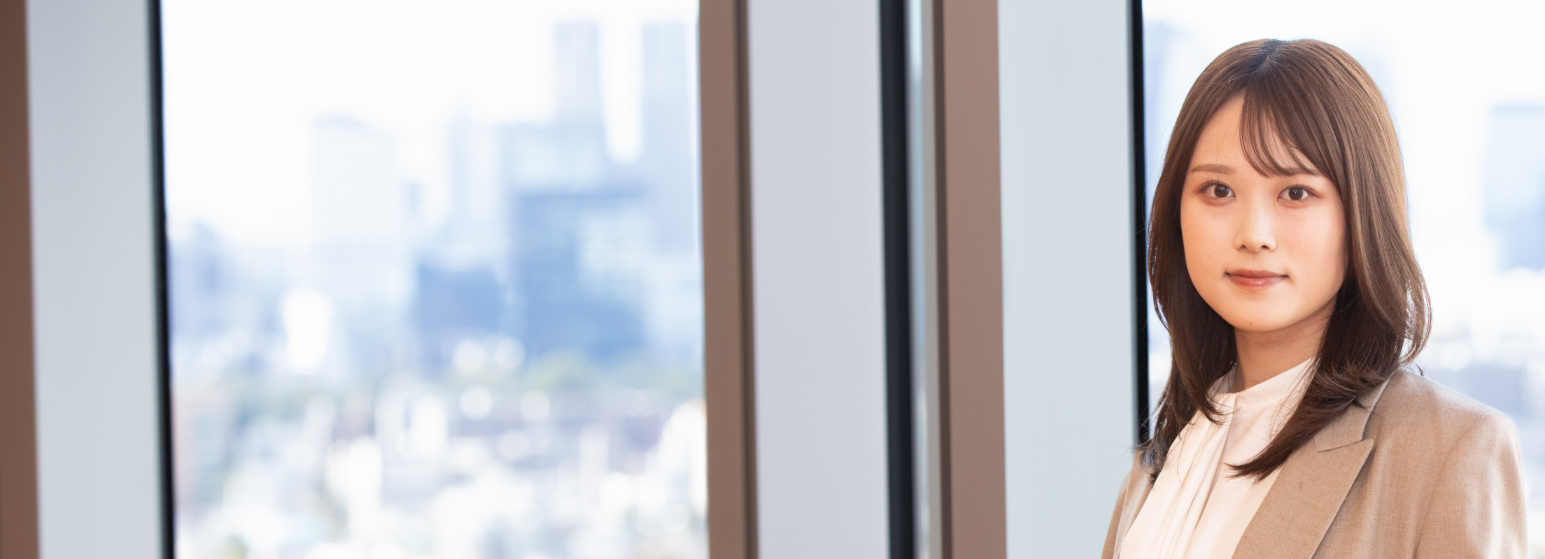 大きな窓の前で上半身が写る杉崎さんのポートレート。白いブラウスとベージュのジャケットを身につけ、落ち着いた表情でカメラを見つめている。背景には都心の高層ビル群がぼんやりと映り、明るく清潔感のある印象。
