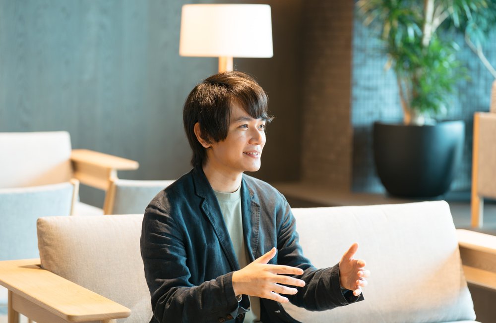 落ち着いた照明のラウンジで、高島さんがソファに座り笑みを浮かべながら語りかけている。
