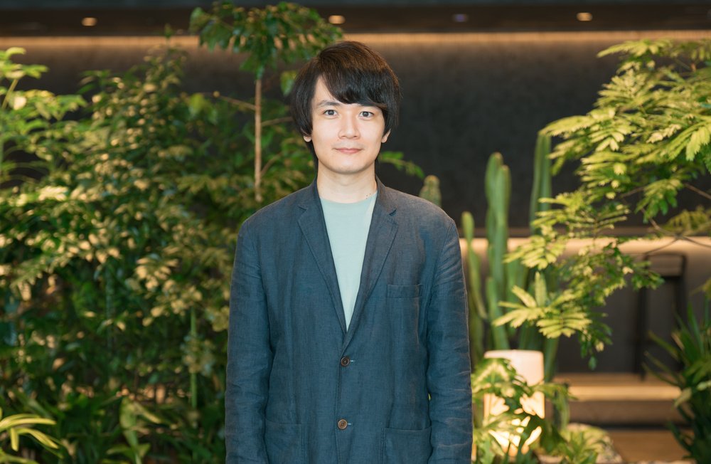 観葉植物やサボテンが並ぶ緑豊かな背景の前で、高島さんが正面を向いて立っている上半身の写真。