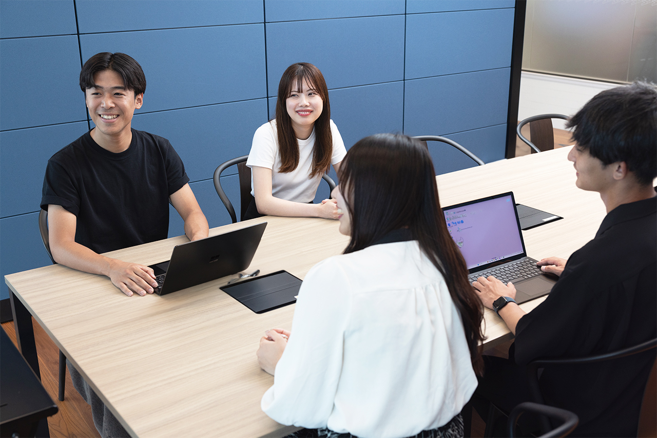 会議室のテーブルで複数の人物が打ち合わせをしている様子。1人がペンを持って資料に書き込みをしており、もう1人が紙の書類を手にしている。机の上にはノートや書類、ペン、グラスなどが並んでいる。左側の白い帯には「ビジネスコンサルタント」「Business Consultant」と書かれている。全体的に自然光が差し込む明るい雰囲気で、落ち着いたビジネスシーンを切り取った構図になっている。