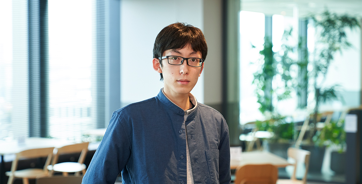 青いシャツを着た眼鏡の男性が、明るい室内で立っています。背景にはテーブルや椅子、観葉植物が見えます。