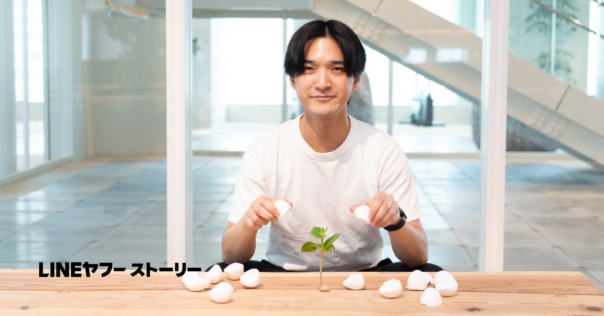 LINEヤフーの中野修平さんが木のテーブルに座り、卵の殻から芽吹いた若葉を前に白い卵を両手で持っている写真。