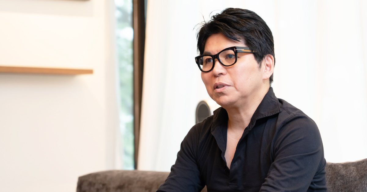 ソファに座り、身ぶりを交えながら話す鎌田さんの写真。窓からの柔らかい光が差し込む室内で、インタビューに応じている。