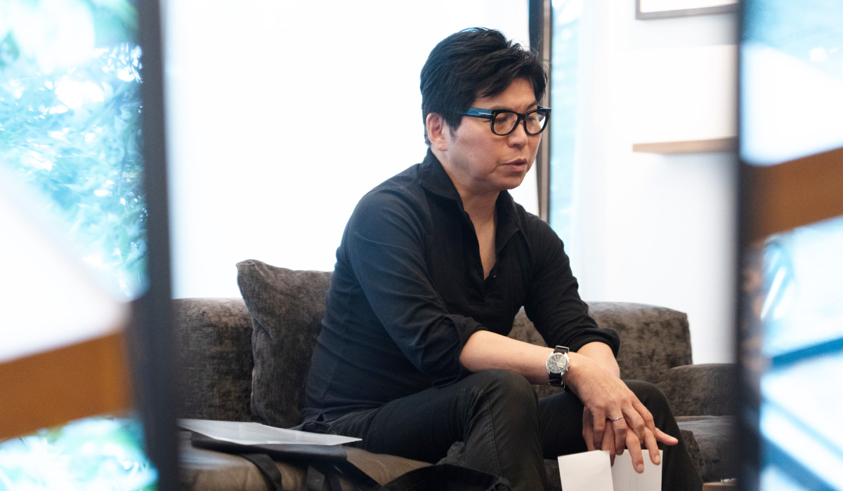 ソファに深く腰掛け、考え込むような表情で話す鎌田さんの写真。手前の鏡越しに撮影され、落ち着いたインタビュー空間が写っている。