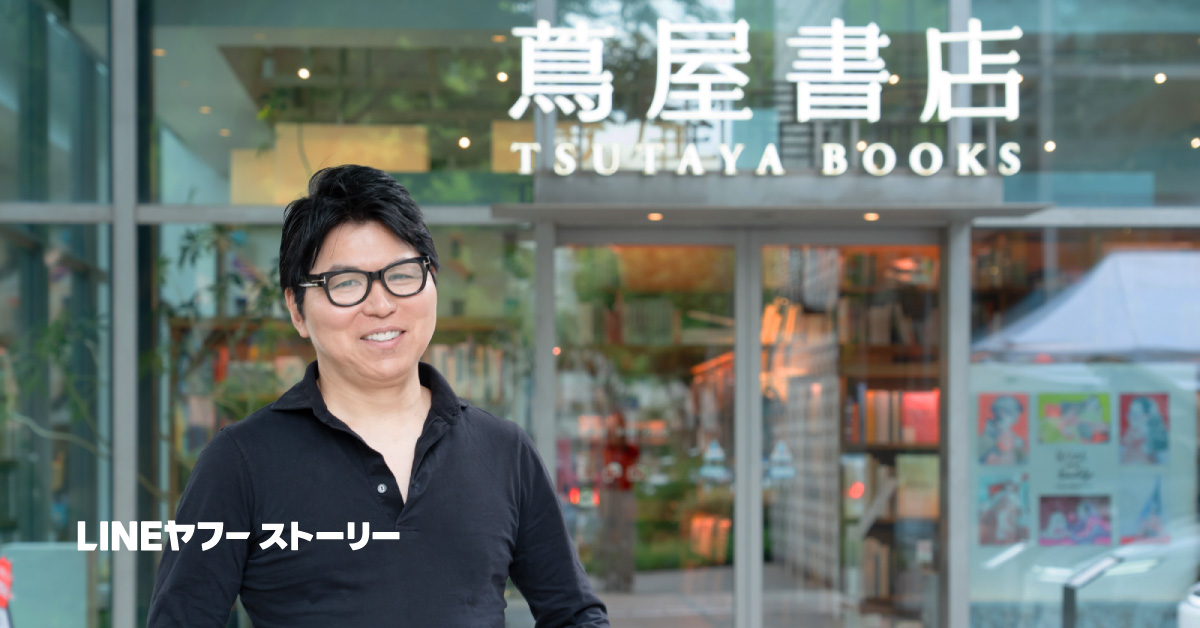 蔦屋書店の入口前に立つCCC戦略店舗開発本部長・鎌田崇裕さんの写真。黒いシャツ姿で笑顔を見せ、背後には蔦屋書店のロゴとガラス越しの店内が見える。