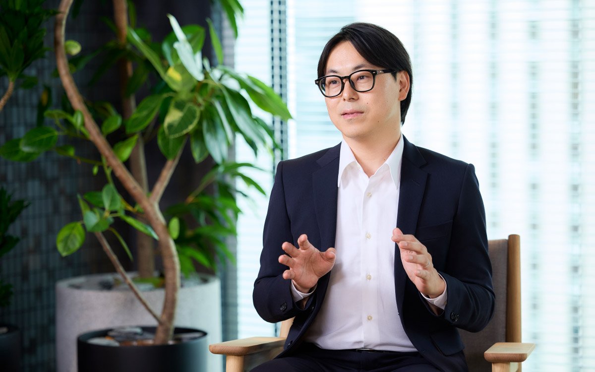 椅子に腰掛け、横向きで両手を使いながら説明する菅谷さんの写真。スタンドライトのある室内で、真剣な表情で語っている。