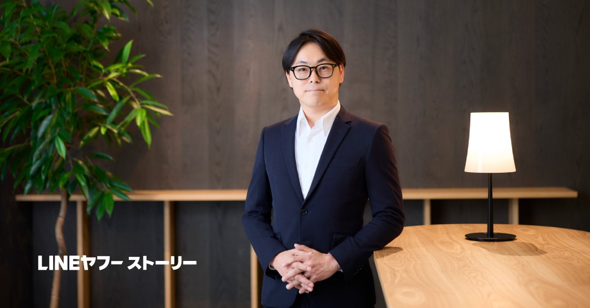 会議室の木のテーブル横に立つLINEヤフーCDO・菅谷聡さんの写真。ネイビーのスーツ姿で正面を見つめ、背後には観葉植物とスタンドライトがある。