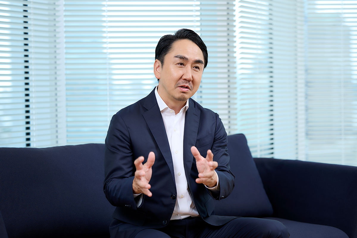 ネイビースーツの男性、出澤がソファに座り、両手を広げて語りかけるように話している様子。背景はブラインドのある窓