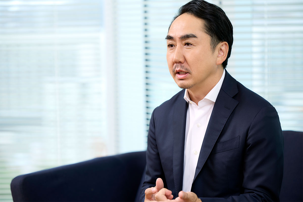 スーツ姿の出澤がソファに腰掛け、体を少し前に傾けながら両手を組むようにして説明している姿。背景には白いブラインドがある
