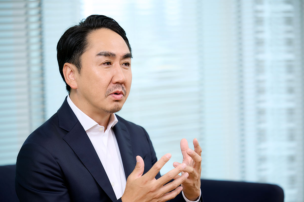 ネイビースーツを着た出澤が体を横に向け、両手を掲げて強調するように話している様子。表情は真剣で、背景はブラインドのある窓