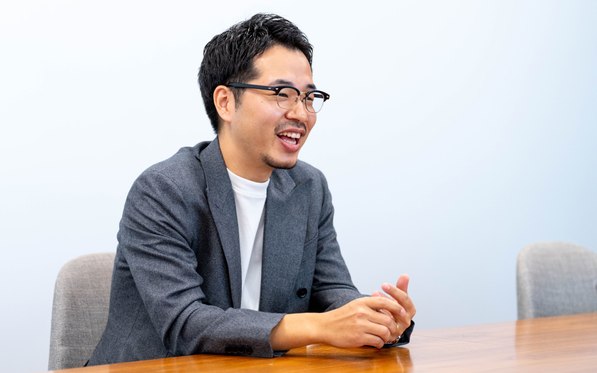 二木のバストショット。木製のテーブルに両手を軽く組むように置きながら、横向きに座って笑顔で会話している様子。視線はやや右方向に向けられ、リラックスした雰囲気。