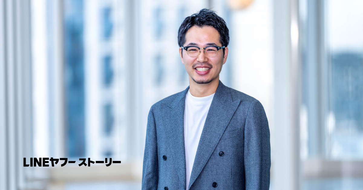 二木のバストショットです。グレーのダブルブレストのジャケットに白いTシャツを合わせた装いで、正面を向いてにこやかに微笑んでいる。背景は明るい室内で、窓越しにぼかされた建物が見える落ち着いた雰囲気