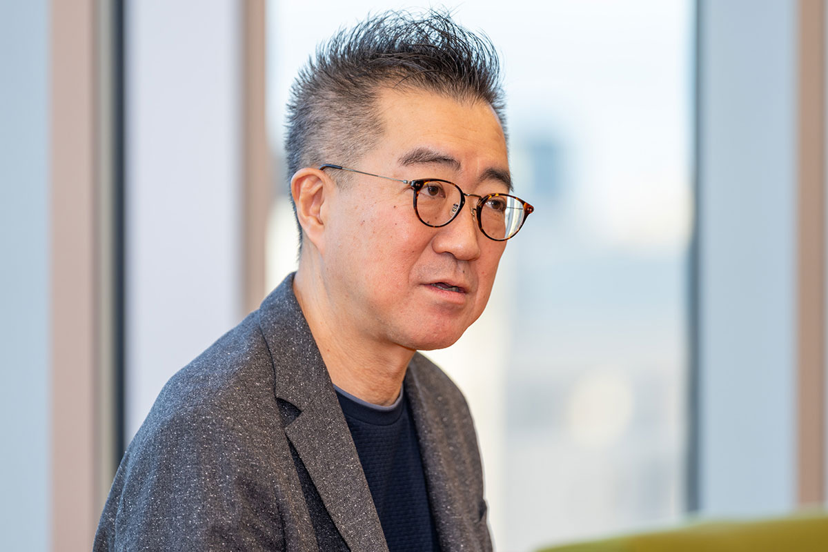 メガネをかけた兵頭CEOが窓際で真剣な表情で話しています。背景には都市の景色があります