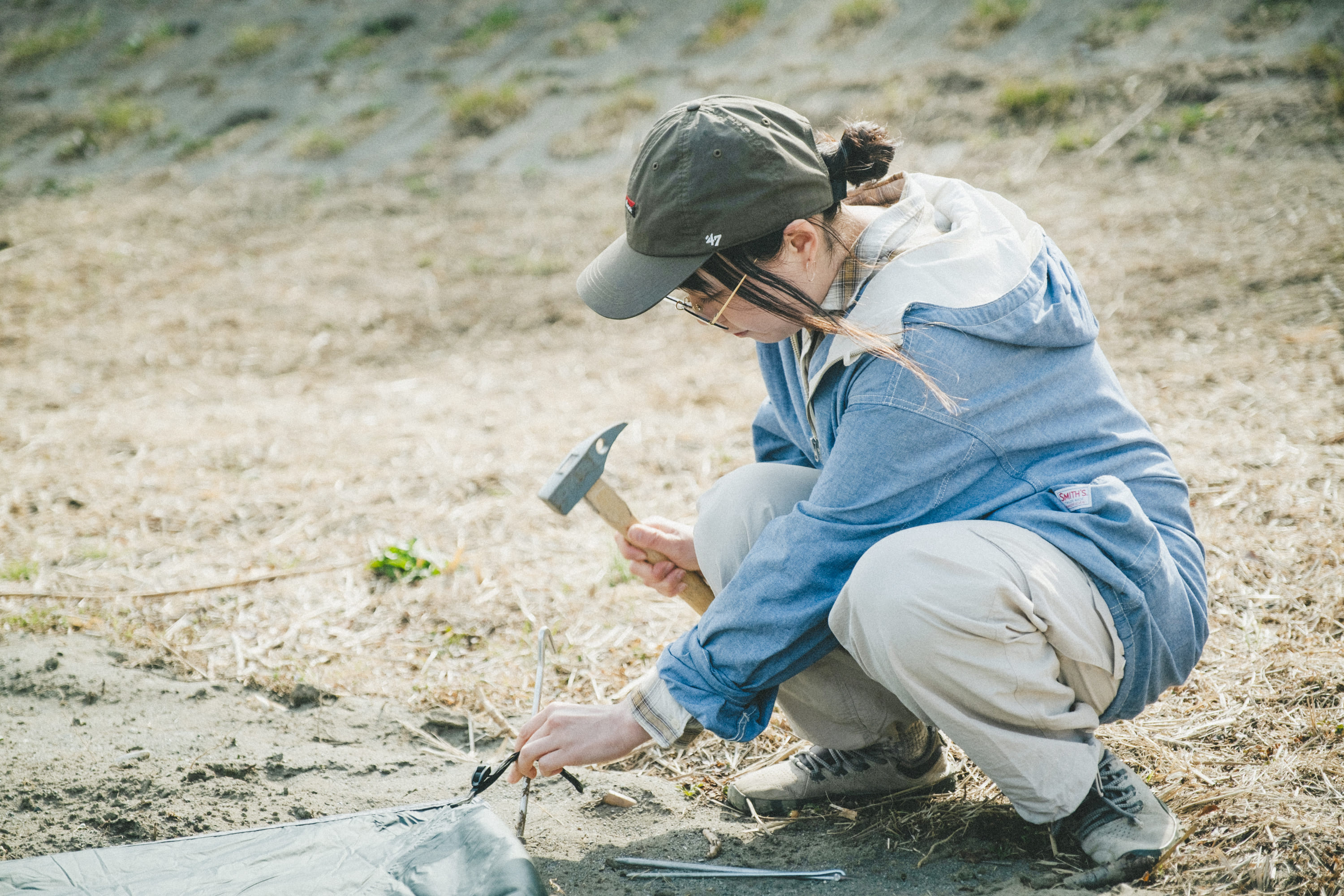 屋外の地面にしゃがみ込み、こいしさんがハンマーでテントのペグを打ち込んでいる写真。
