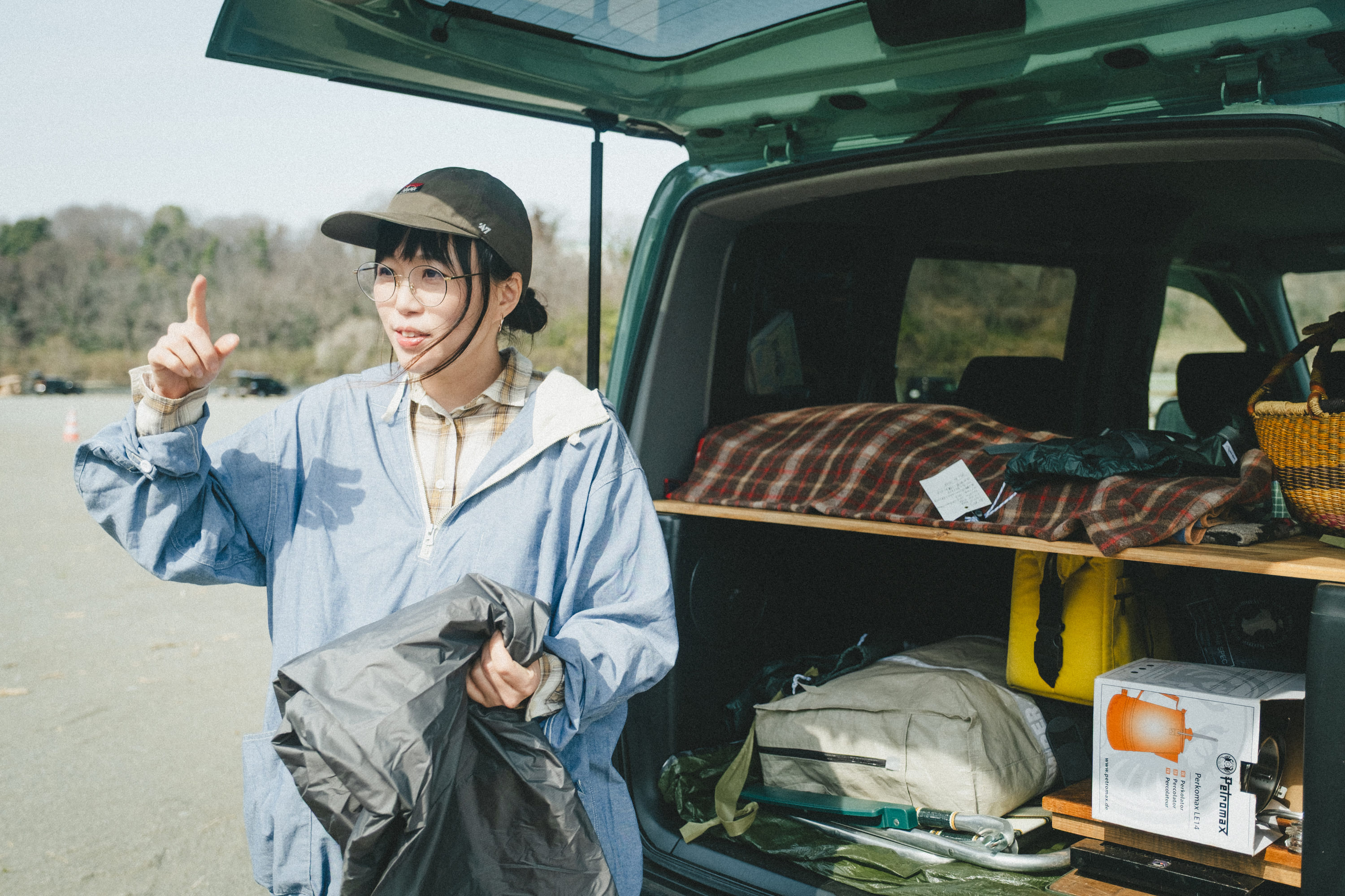 車の後部ハッチを開けた前で、こいしさんが立ちながら左手の人差し指を上げている写真。車内には木製の棚が設置され、チェック柄のブランケットや黄色のバッグ、カーキ色の収納袋、箱入りのランタンなどのキャンプ道具が整然と積まれている。